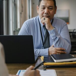 Serious mature ethnic entrepreneur sitting at wooden table with documents and laptop and thinking while crop anonymous colleague taking notes on clipboard