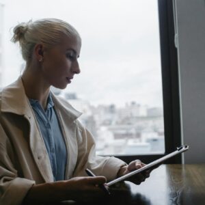 Side view of young concentrated female wearing white coat sitting at wooden desk in office and checking reports on clipboard